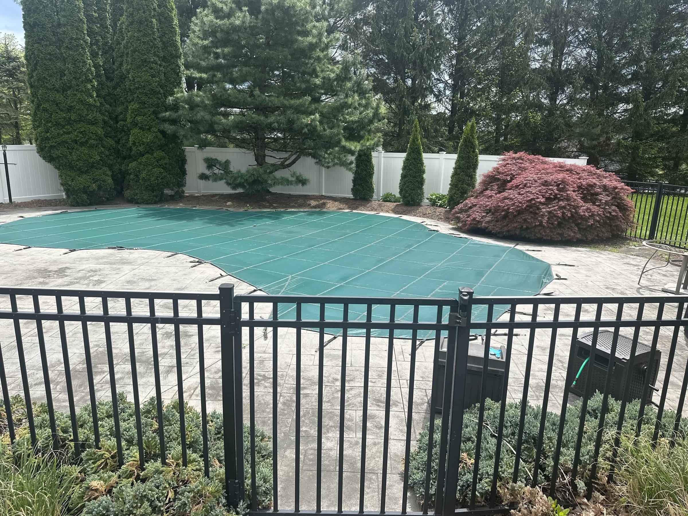 Black aluminum pool fence on a stamped concrete patio around a green safety-covered inground pool with white vinyl fence behind