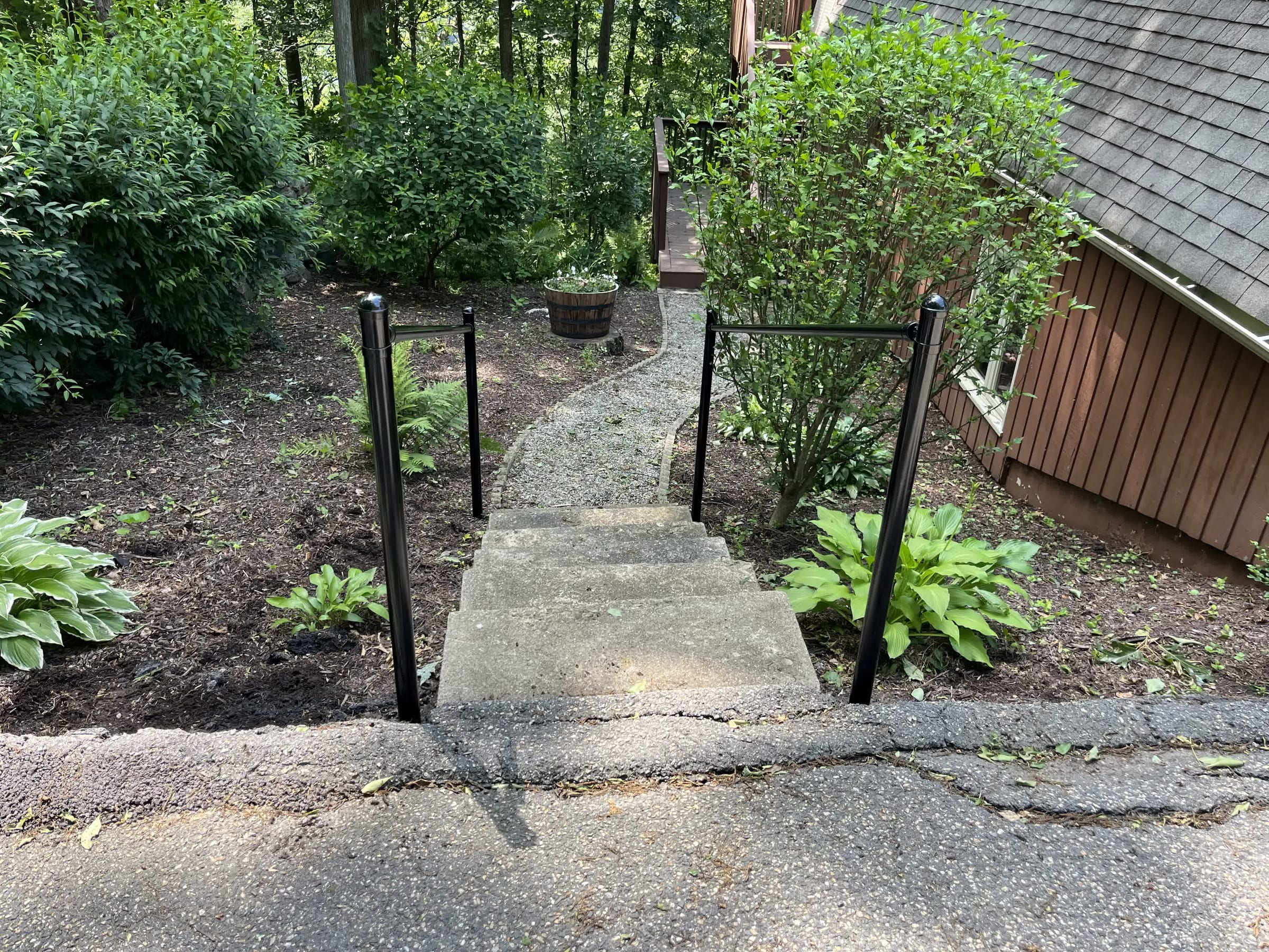 Two black metal handrails flanking concrete steps that lead to a gravel garden path among hostas and shrubs