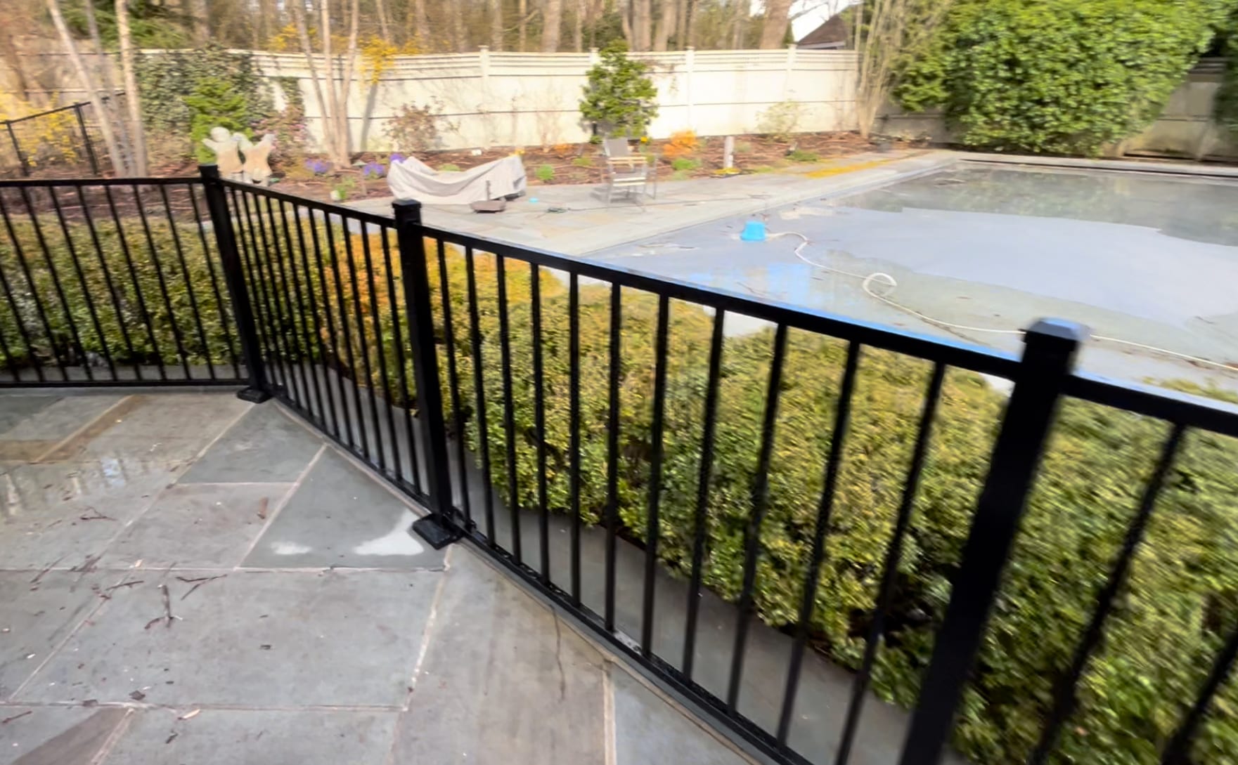 Black aluminum railing along the edge of a bluestone patio beside a covered in-ground pool and hedges