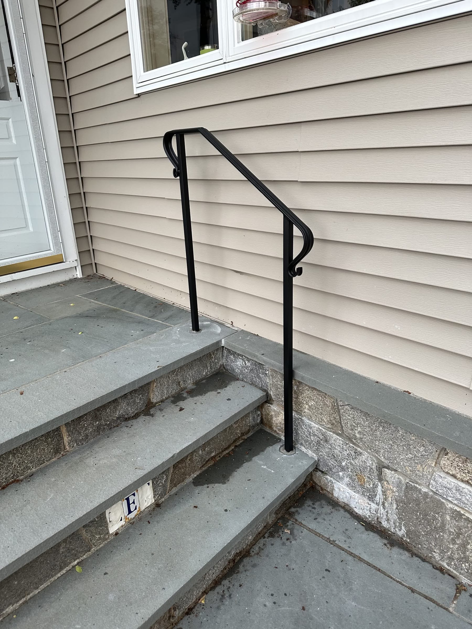 Black ornamental handrail with scroll ends on bluestone entry steps against tan clapboard siding