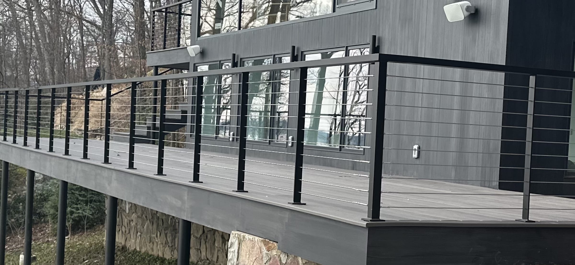 Black metal posts with horizontal stainless cable railing along an elevated composite deck on a modern gray house