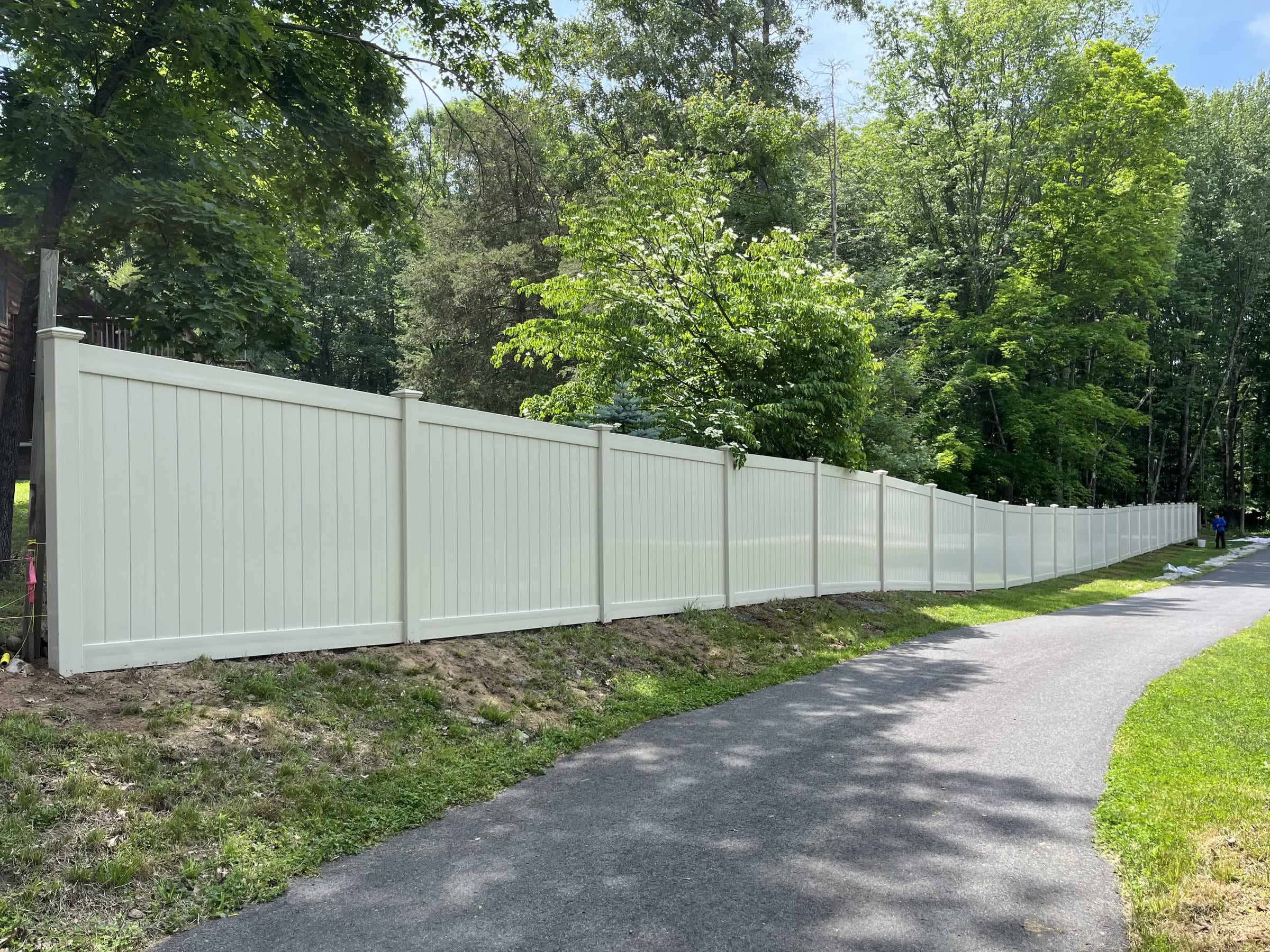 Long cream-colored vinyl privacy fence running downhill beside a paved driveway with dense green trees behind.
