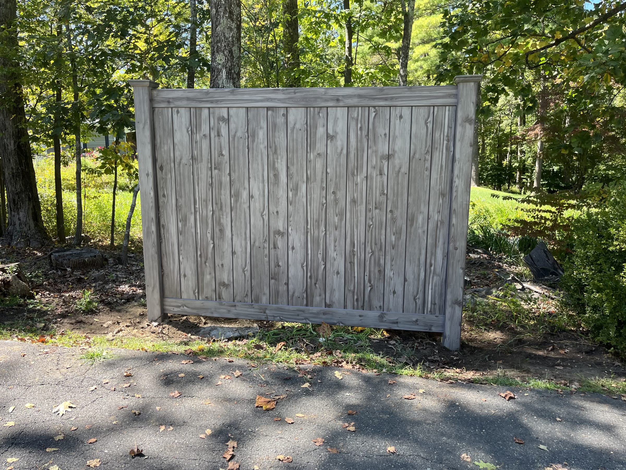 Single gray woodgrain vinyl privacy fence panel with capped posts beside a driveway with sunlit woods behind.