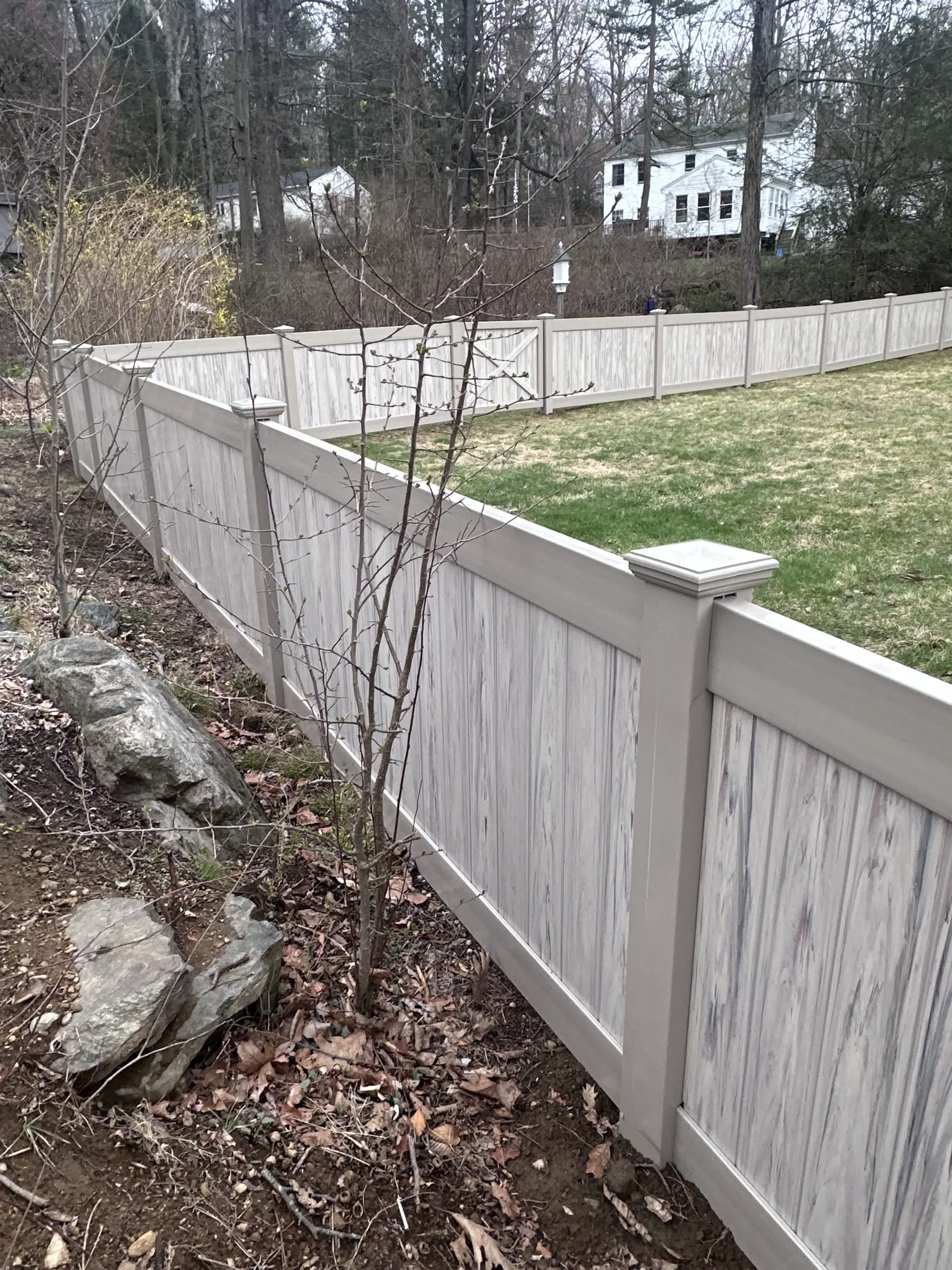 Gray woodgrain vinyl privacy fence running along a sloped wooded yard with a rock border and young tree.