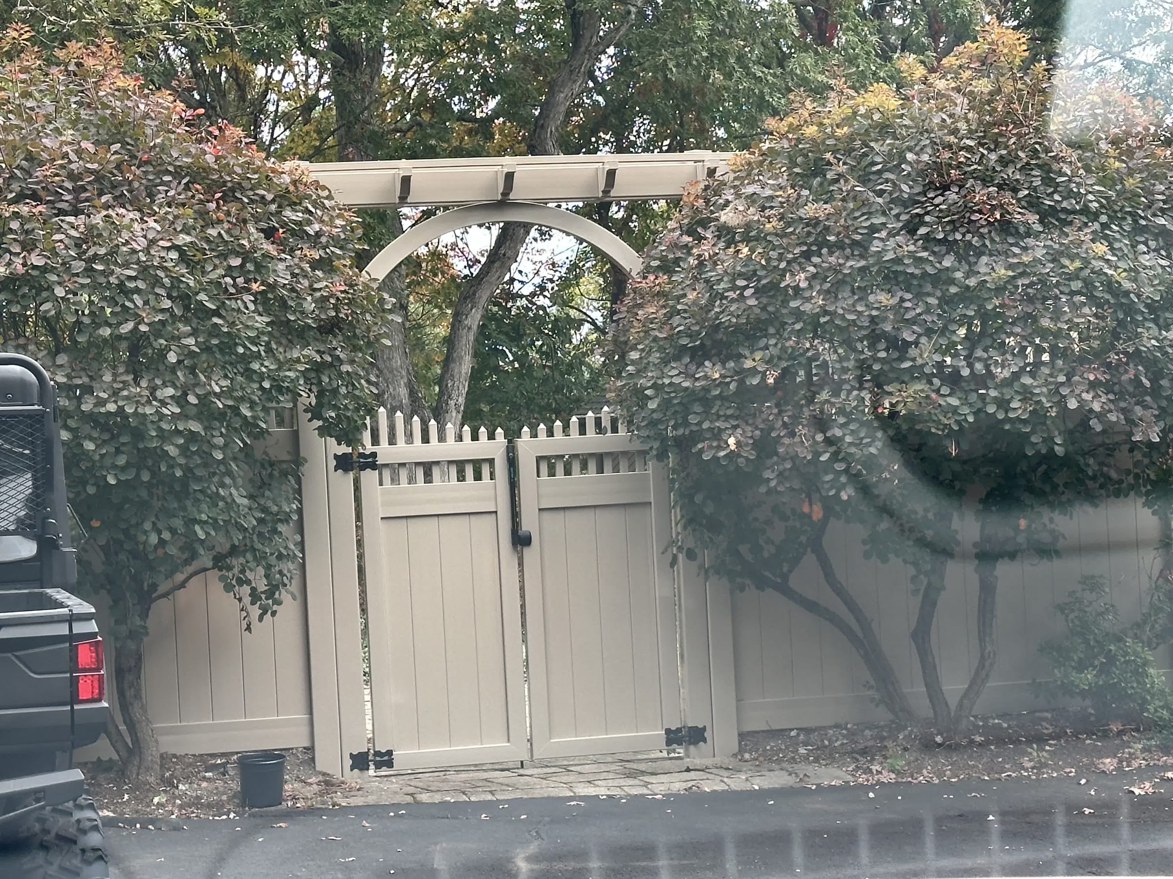 Tan vinyl privacy fence with an arched arbor over a double gate, framed by manicured shrubs at a driveway edge.