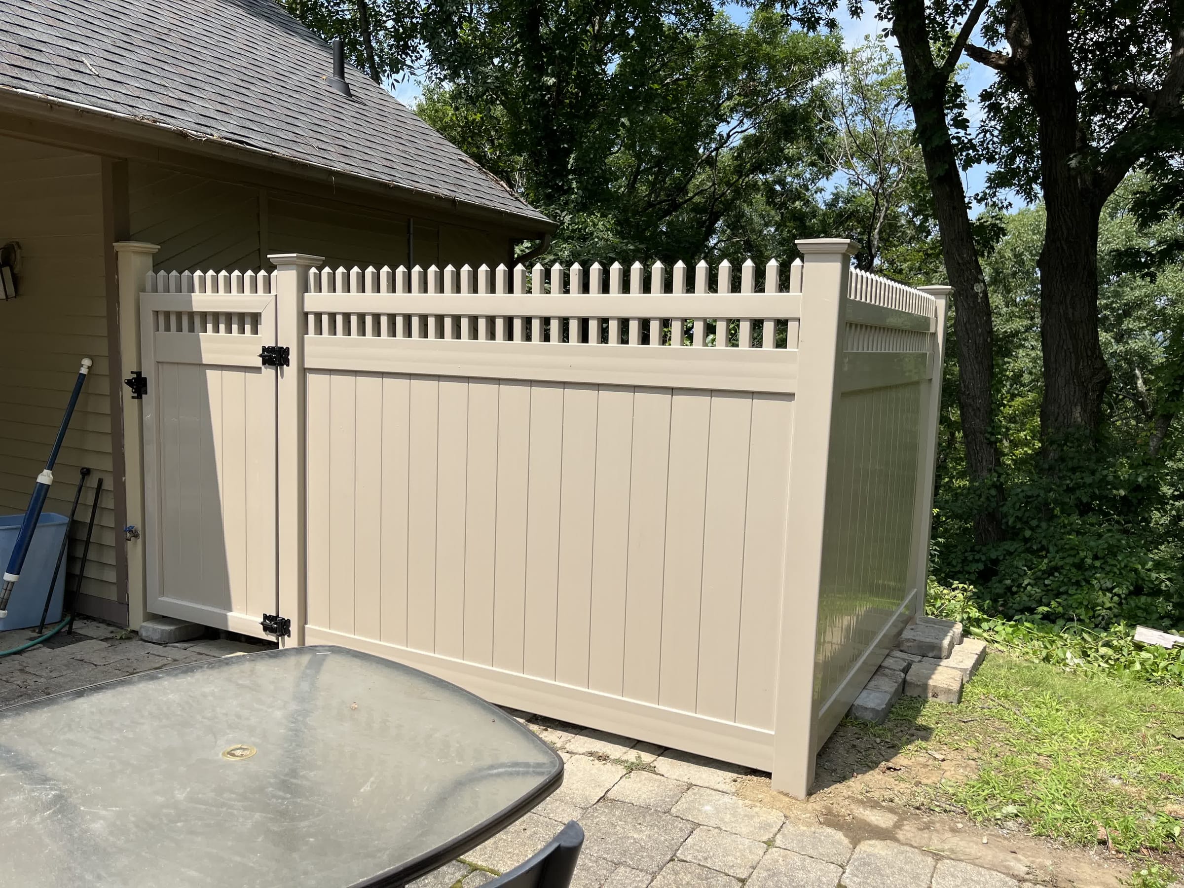 Tan vinyl privacy fence with open picket topper and a gate enclosing a paver patio beside a house and trees.