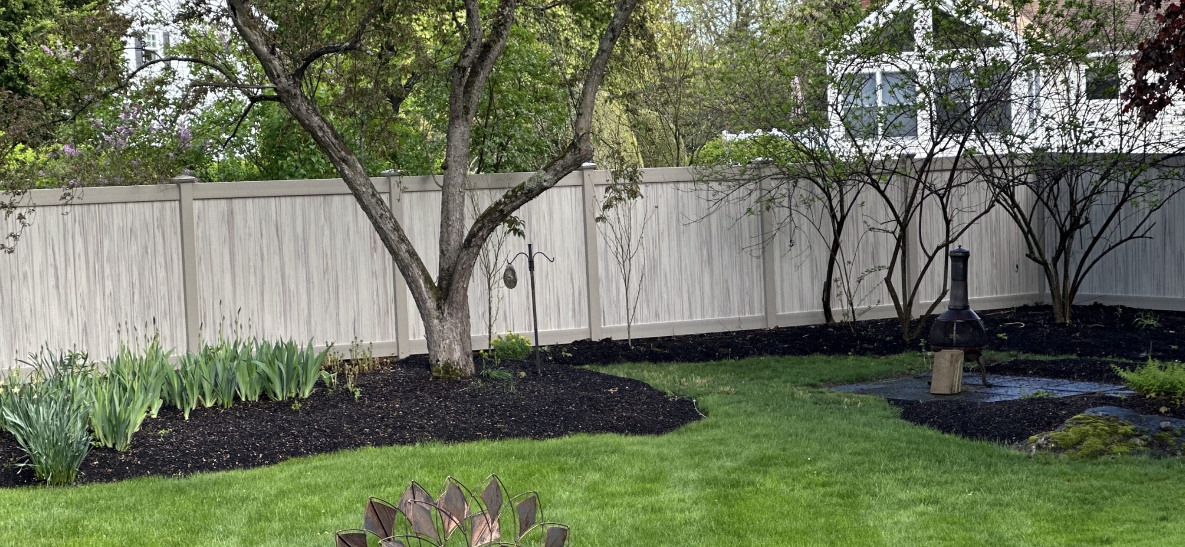 Tan woodgrain vinyl privacy fence bordering a landscaped backyard with mulch beds, trees and a chiminea.