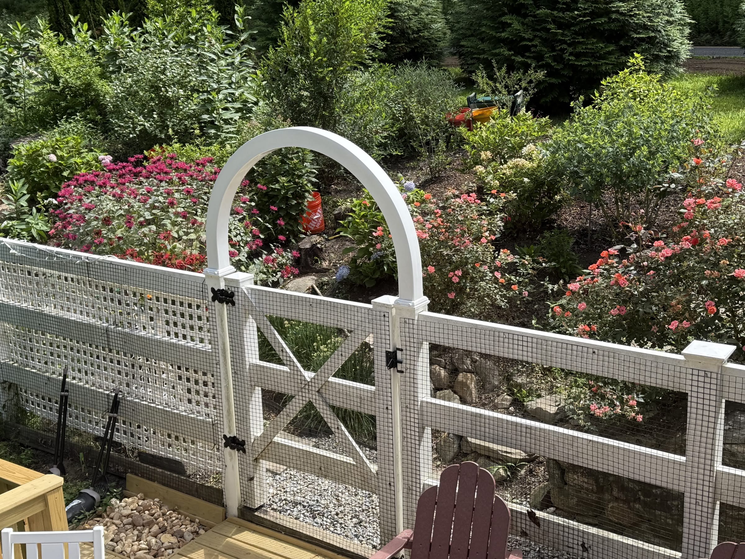 White square-lattice garden fence with an arched arbor gate and wire mesh, bordering a flowering perennial garden.