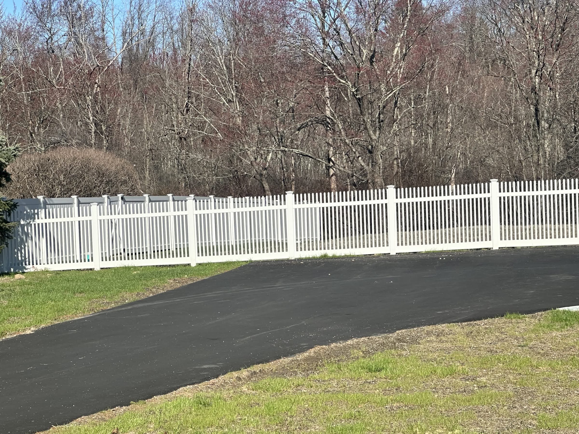 White vinyl picket fence running along a paved driveway edge with bare early-spring trees behind under blue sky.