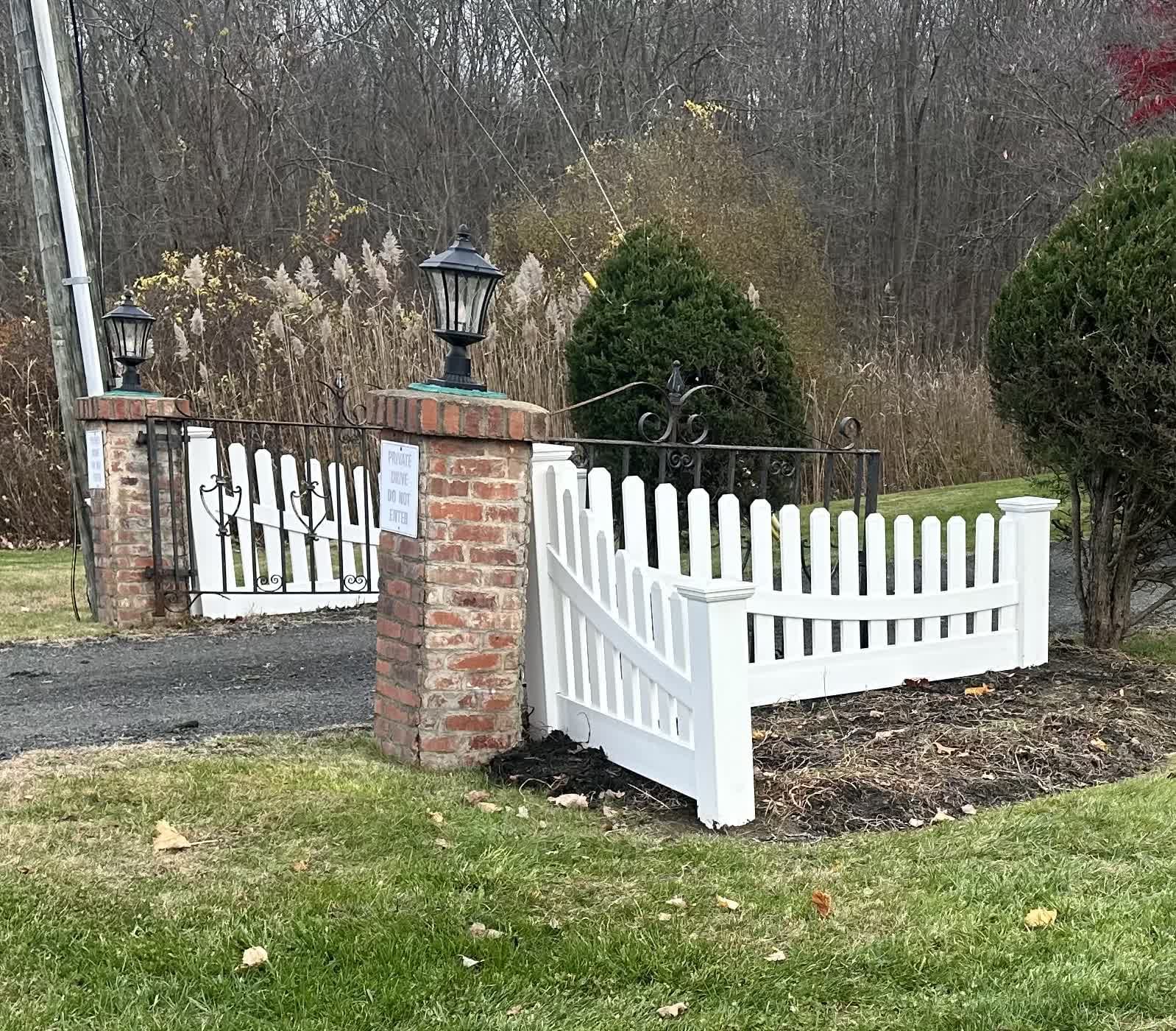 White vinyl picket fence adjoining brick pillars with lantern caps and an iron gate at a driveway entrance in fall.