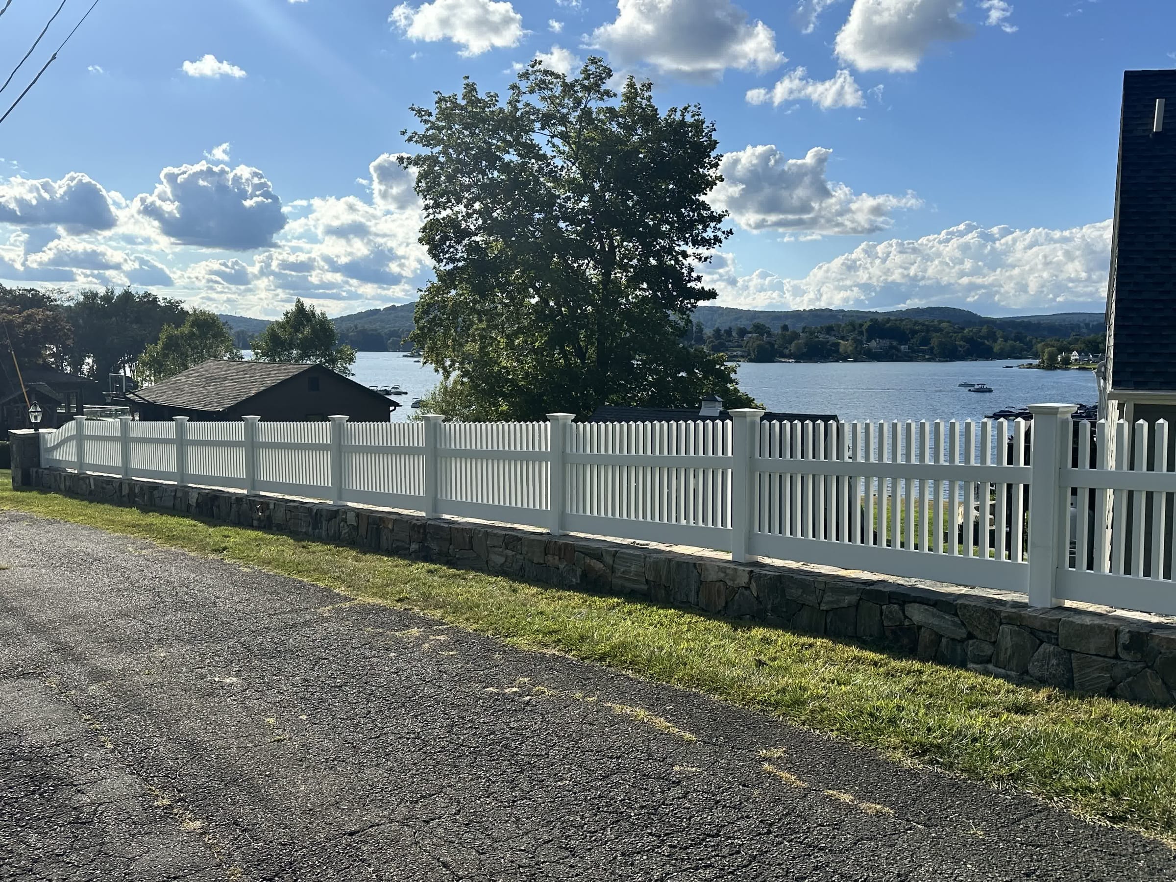 White vinyl picket fence atop a stone retaining wall along a driveway overlooking a lake with hills and boats.