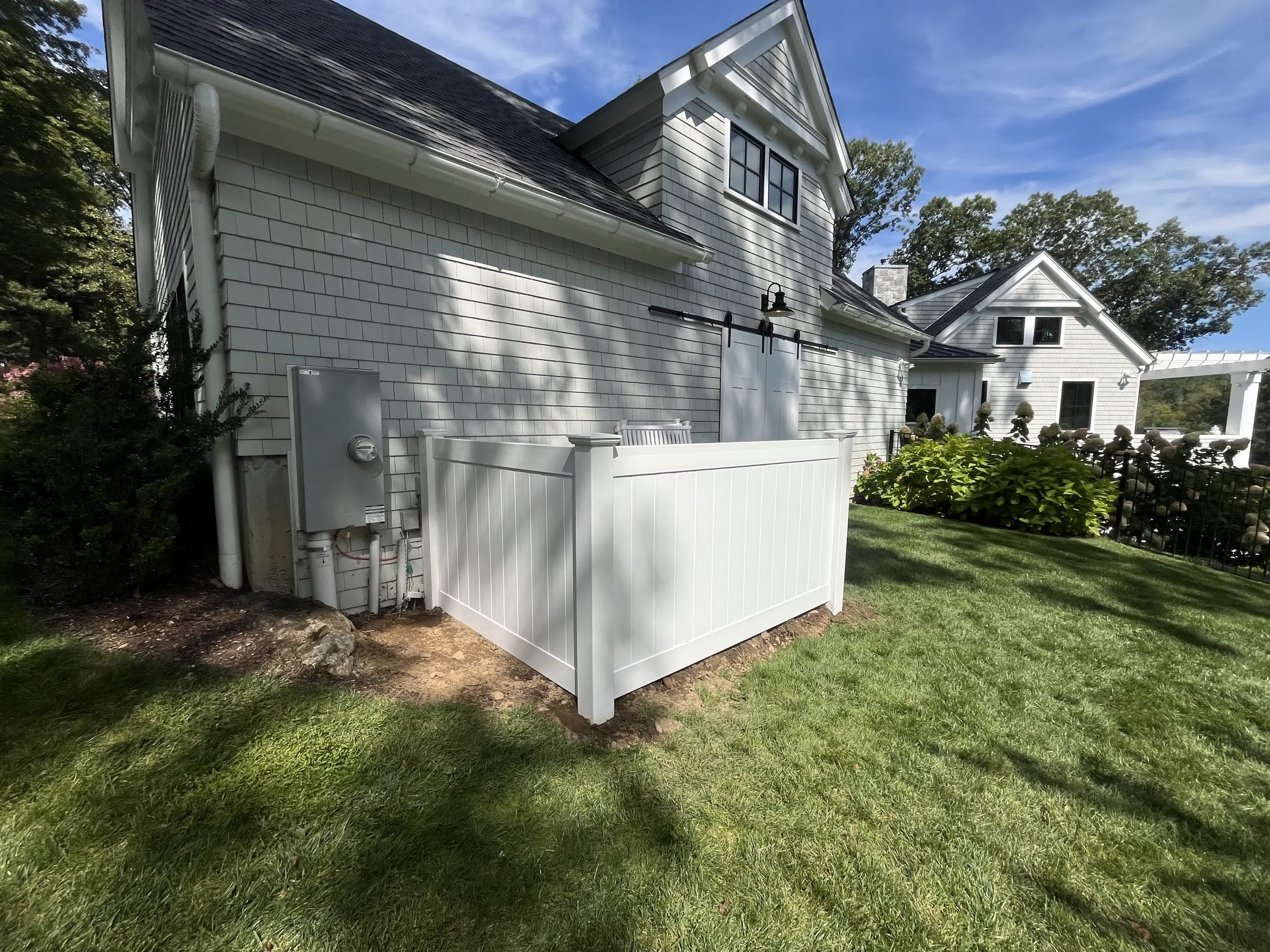 White vinyl privacy enclosure hiding utility meters and equipment at the side of a large shingled home with blue sky.