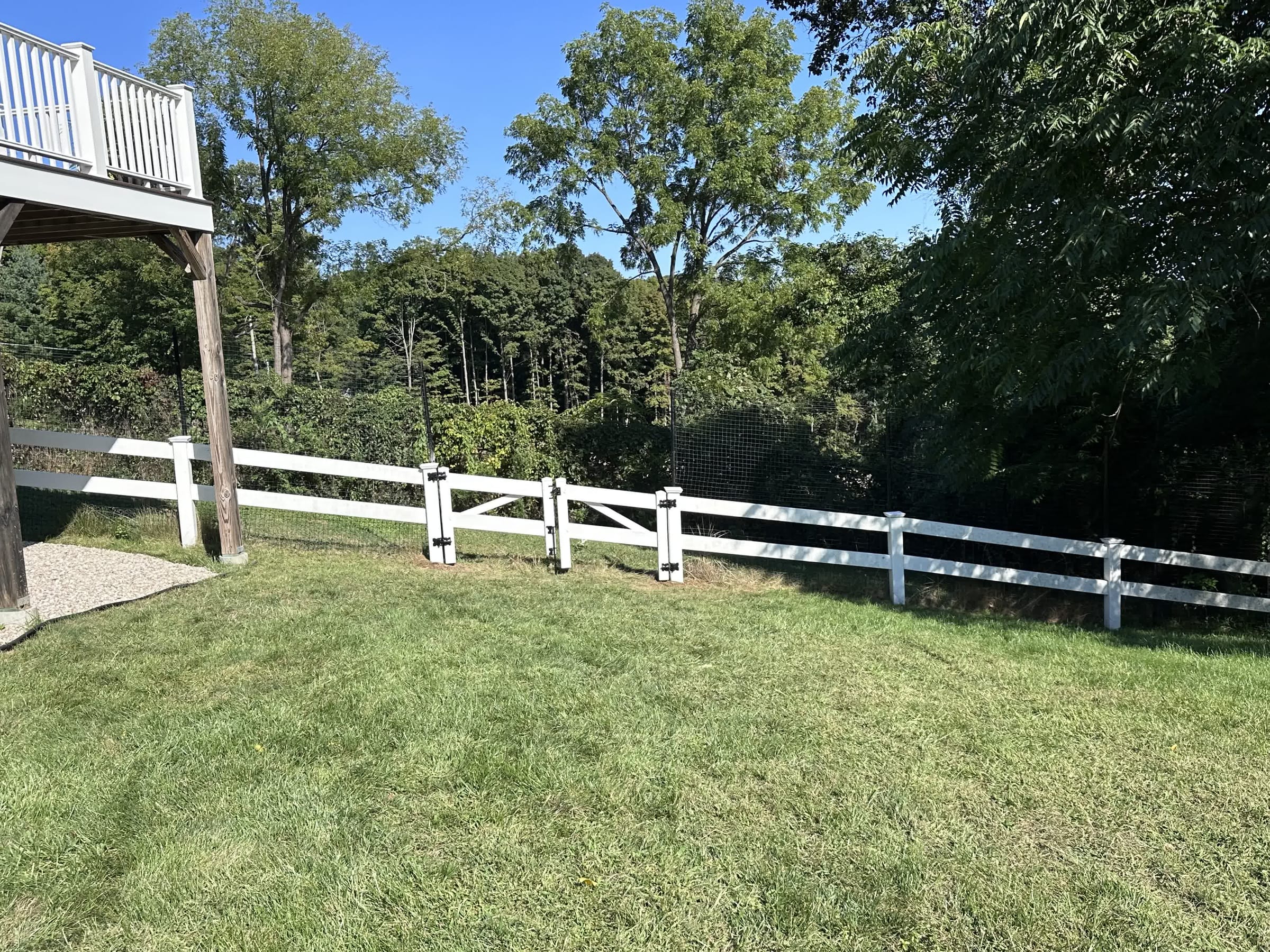 White vinyl two-rail ranch fence with a gate and wire mesh backing along a mowed lawn beside a raised deck and woods.