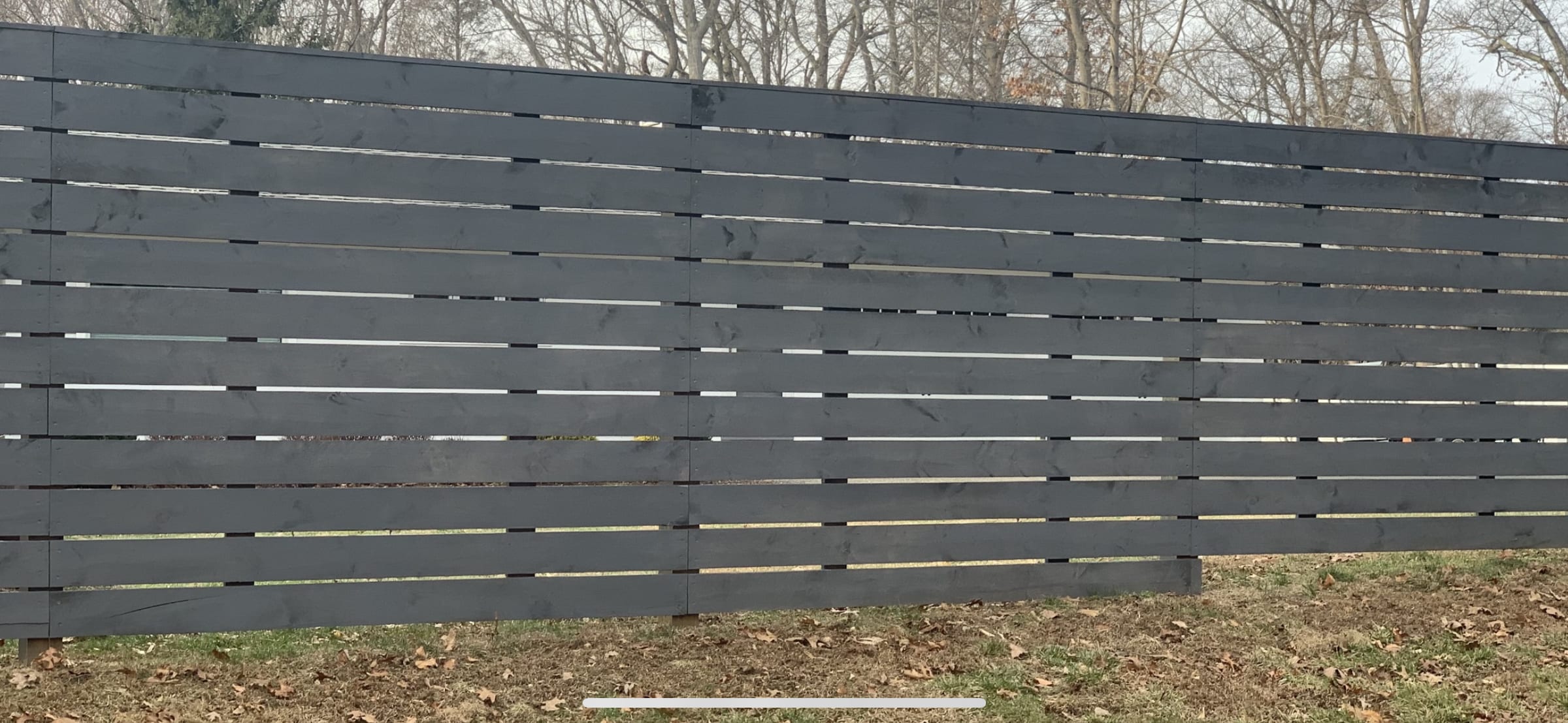 Black-stained horizontal slat semi-privacy fence with narrow gaps set on a grassy yard edge in winter