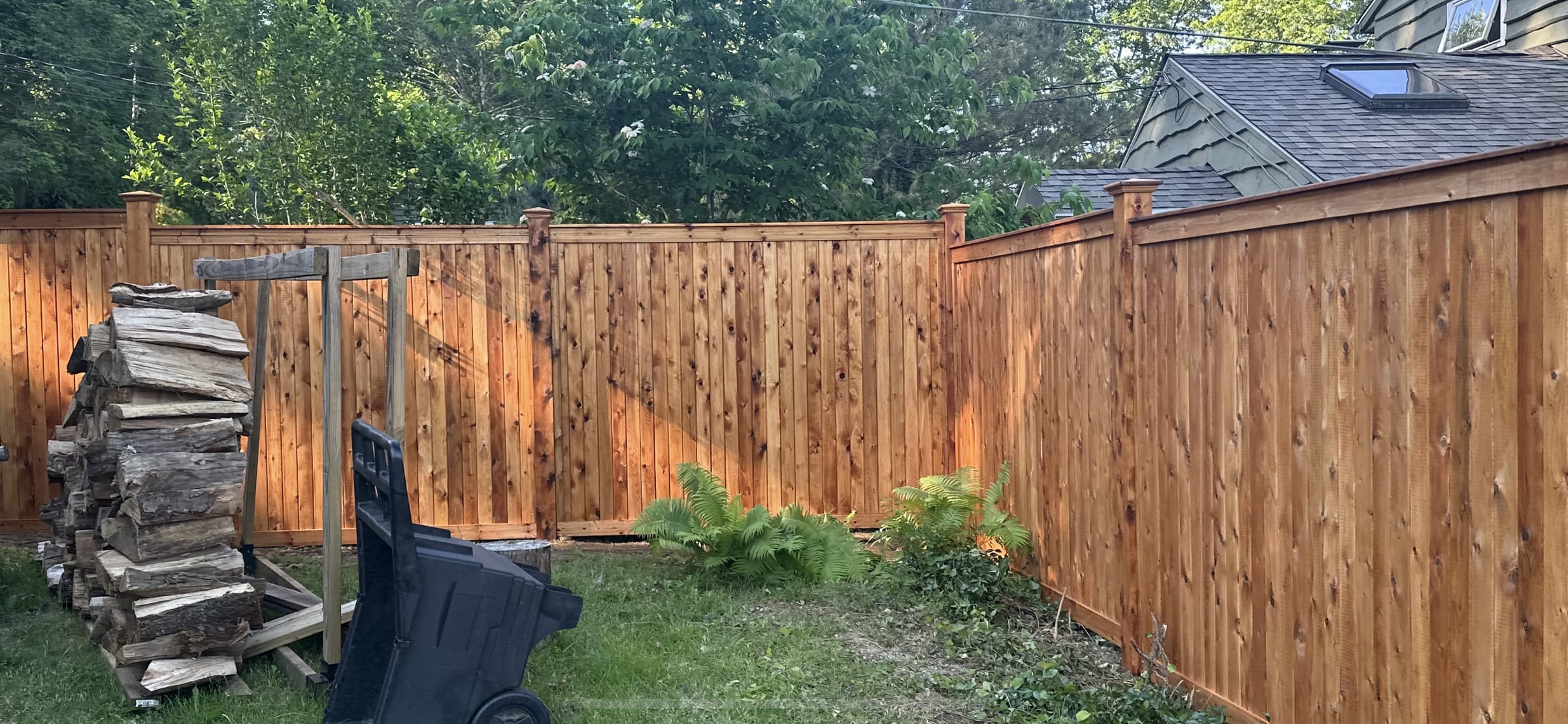 Solid cedar board privacy fence with post caps enclosing a backyard beside a house, firewood stacked nearby