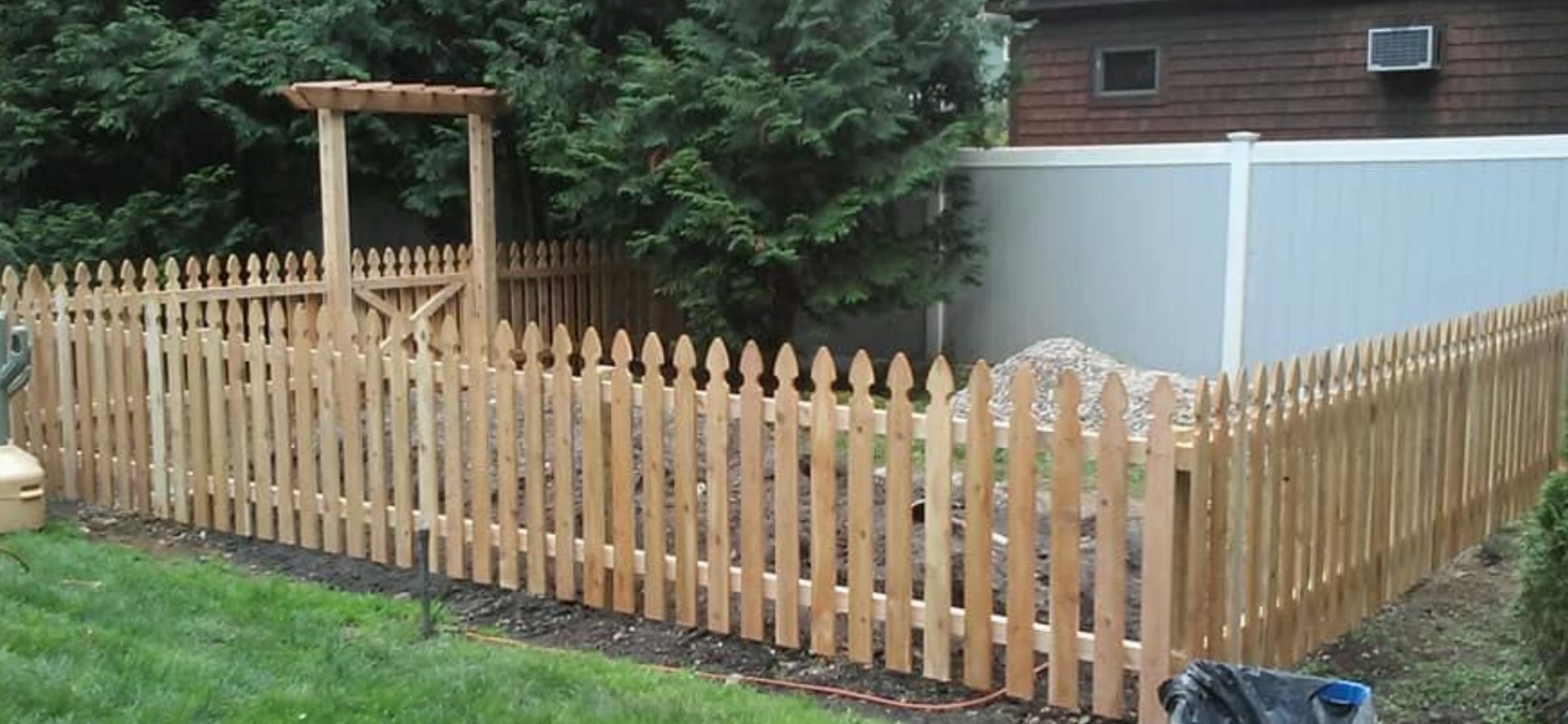 Cedar spaced picket fence with pointed gothic tops and an arbor gateway around a backyard garden bed