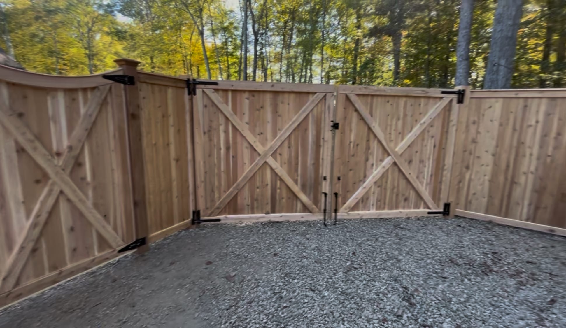 Cedar board privacy fence with a double gate and X-brace backs enclosing a gravel area beside woods