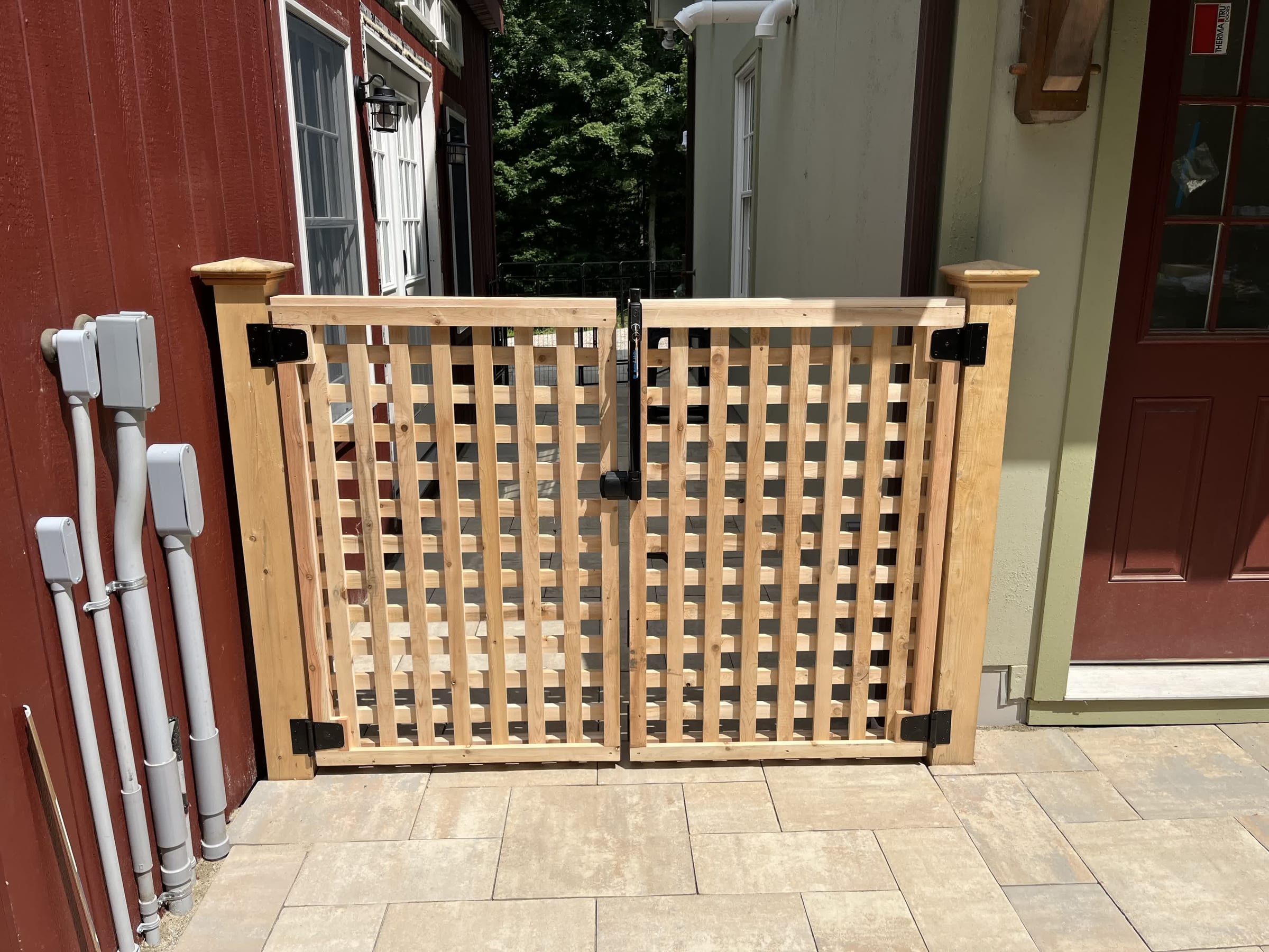 Cedar square-lattice double gate with black hardware set between a red barn and house on a paver patio