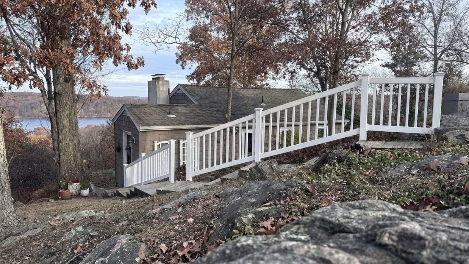 White wooden railing installed on sloped terrain connecting to lakeside house with autumn trees in background