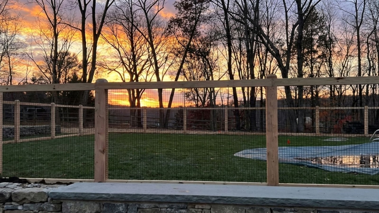 Straight-on view of a wood post and wire mesh fence enclosing a backyard pool with a wooded tree line at sunset