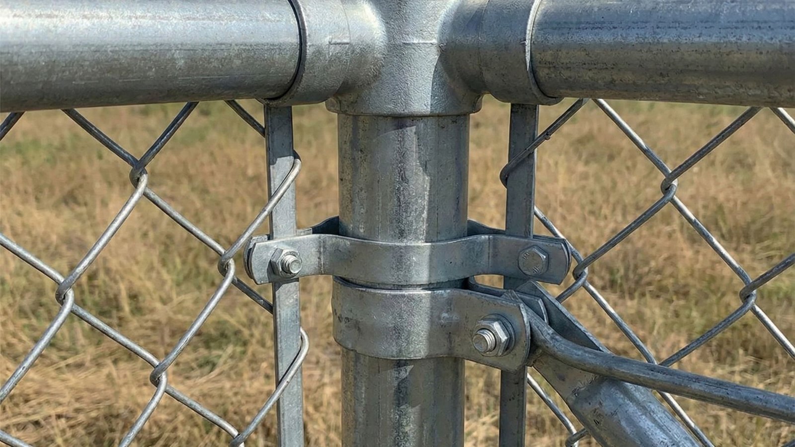 Close-up view of galvanized chain link fence post with metal clamps and hardware securing fence panels to vertical post