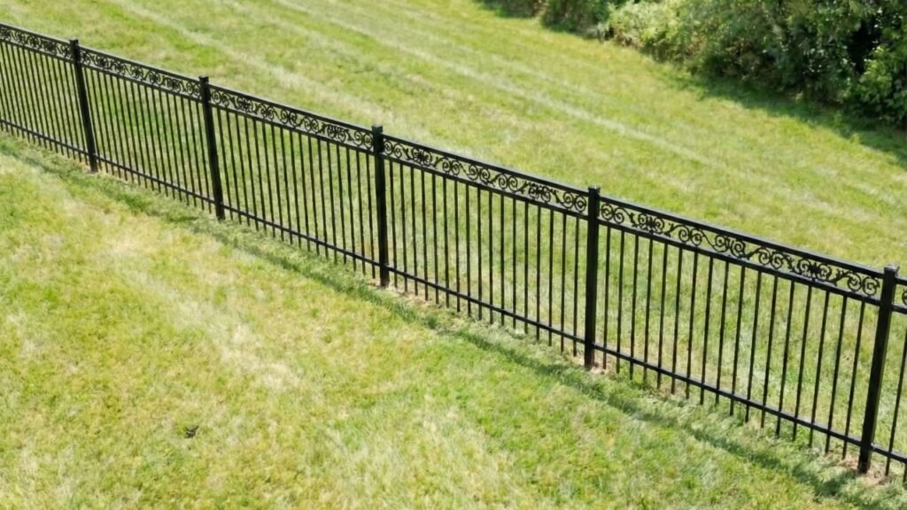 Black ornamental iron fence with decorative scrollwork running across green lawn with trees in background