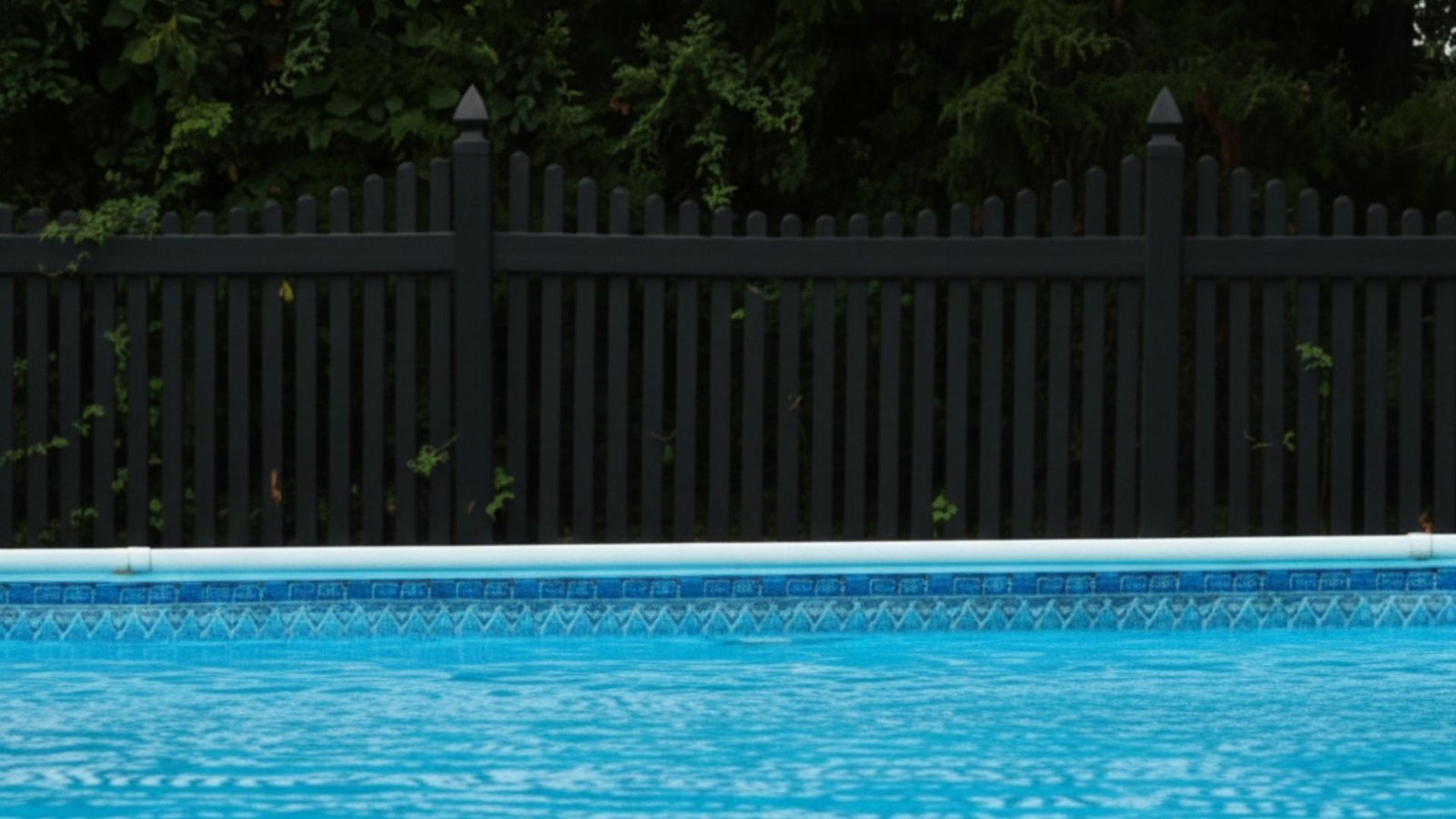 Blue swimming pool with decorative tile border surrounded by black privacy fence and lush green trees