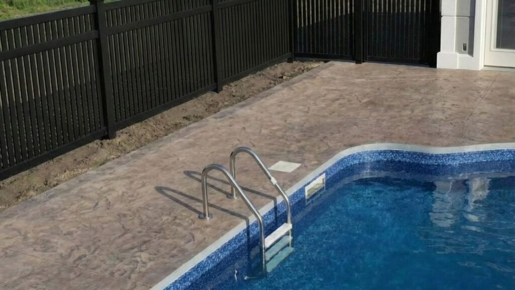Swimming pool with blue tile and stainless steel ladder, surrounded by black privacy fence on stamped concrete deck