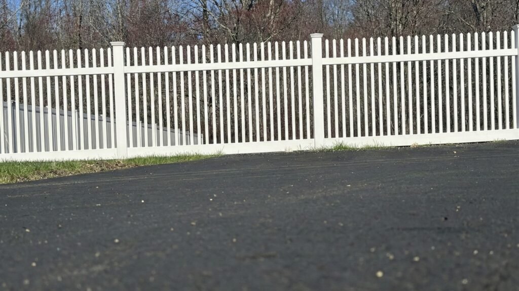White vinyl picket fence with posts along asphalt driveway with bare trees in background during winter season