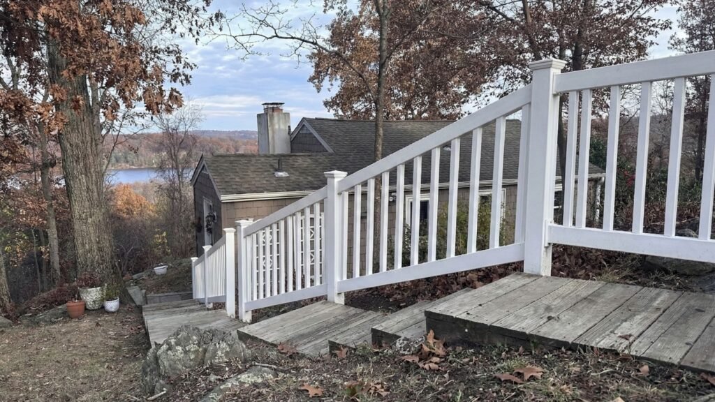 White deck railing installation on wooden deck overlooking lake with autumn foliage and residential home in background