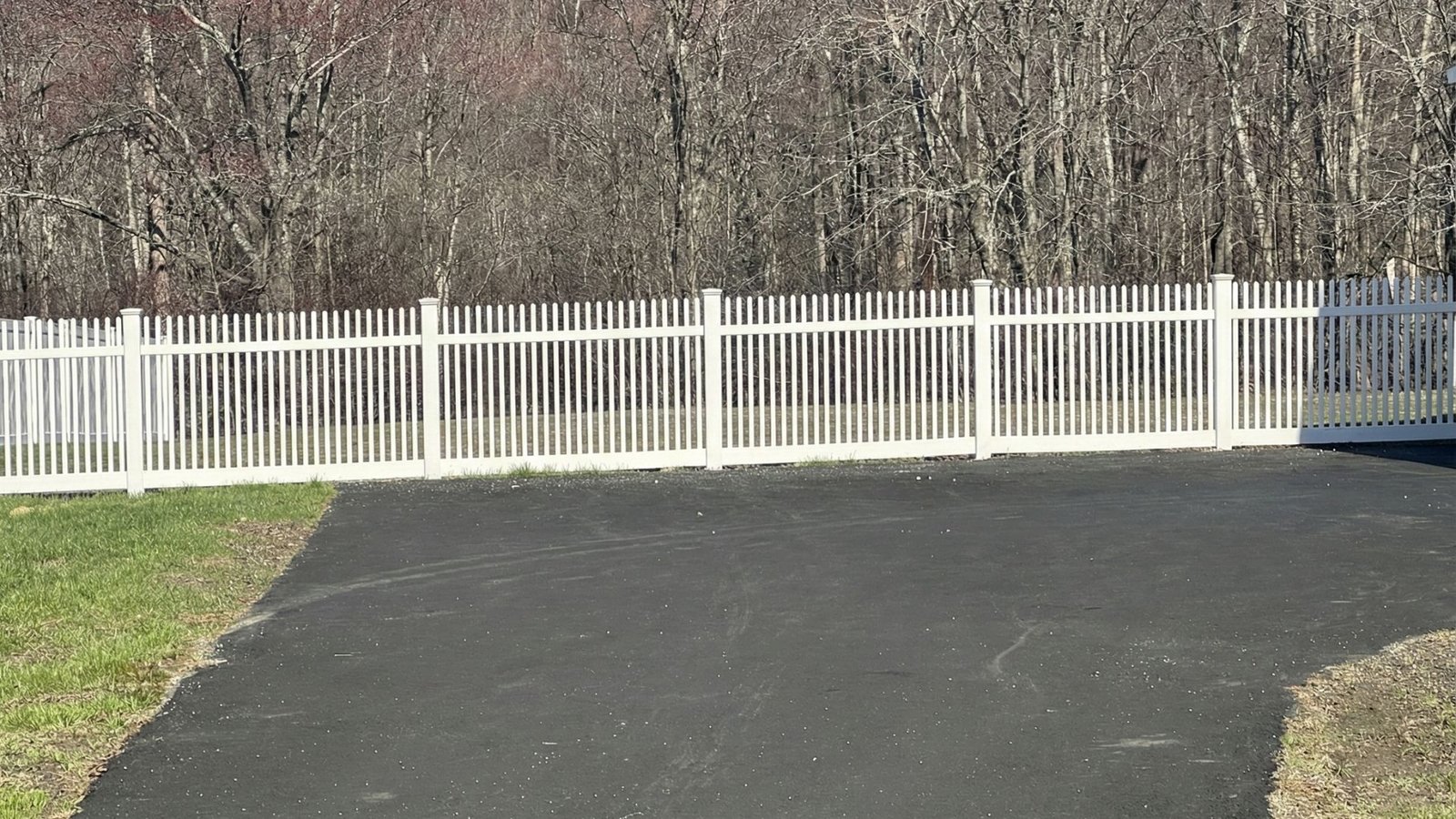 White vinyl picket fence with vertical slats along black asphalt driveway with green lawn and bare trees