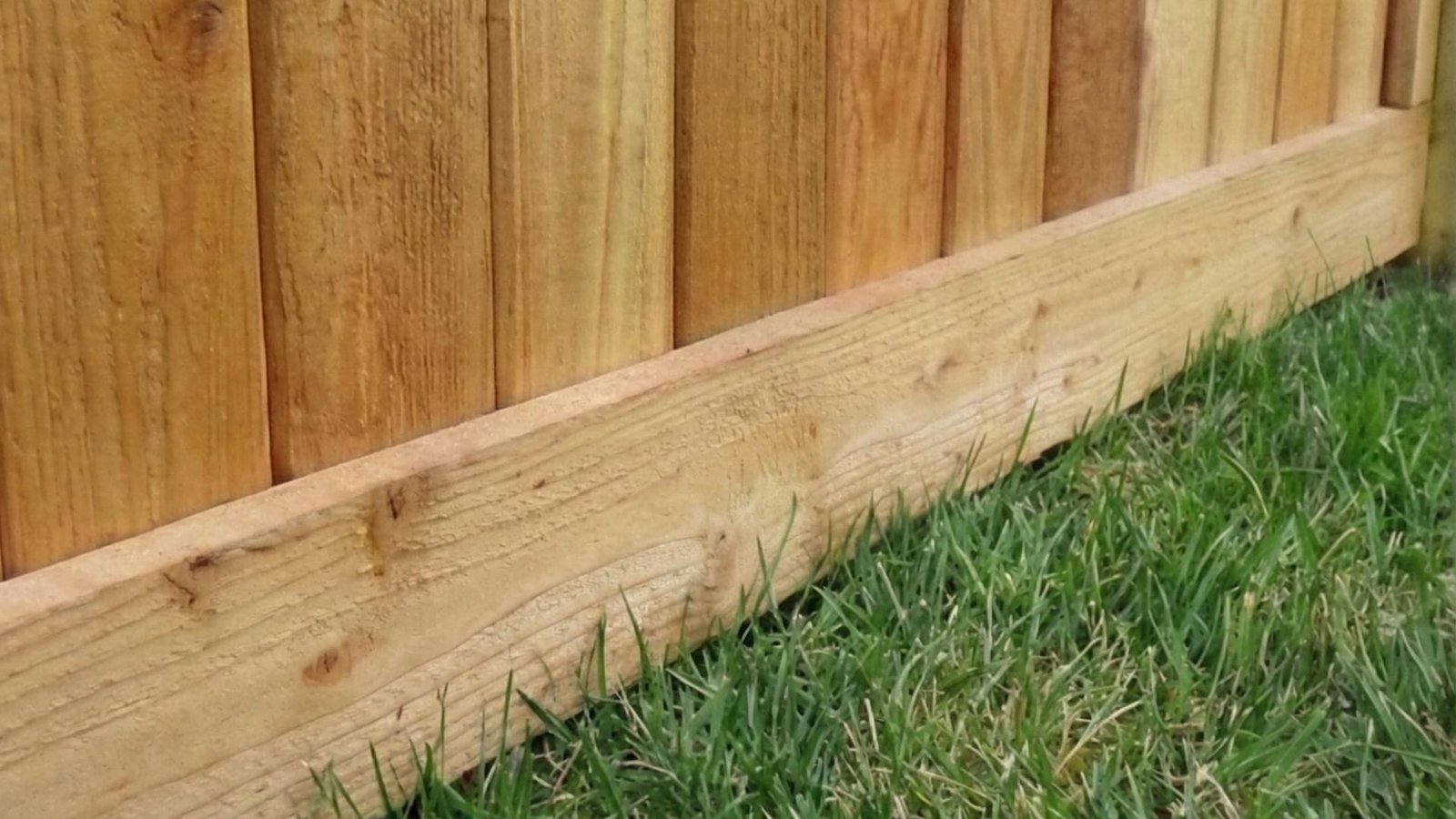 Close-up view of natural wood fence with vertical cedar pickets and horizontal bottom rail meeting green grass lawn