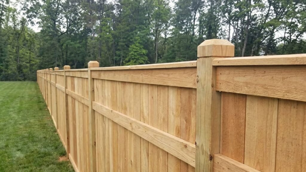 New wooden privacy fence with vertical boards and rounded posts extending across green lawn with trees in background