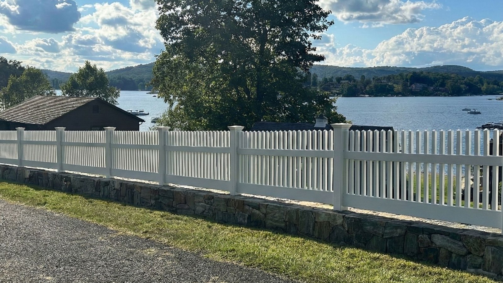 White picket fence on stone retaining wall overlooking lake with trees and cloudy sky in background