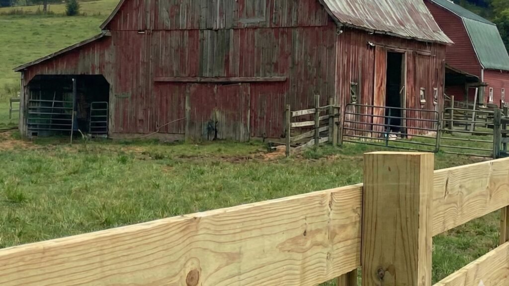 Weathered red wooden barn with metal roof surrounded by wooden rail fencing in rural Amenia NY farmland setting