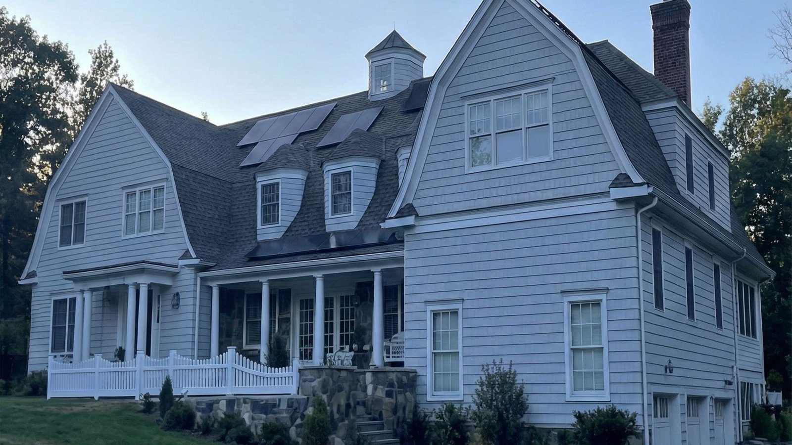 Large white colonial house with stone foundation, front porch, dormers, and solar panels on roof